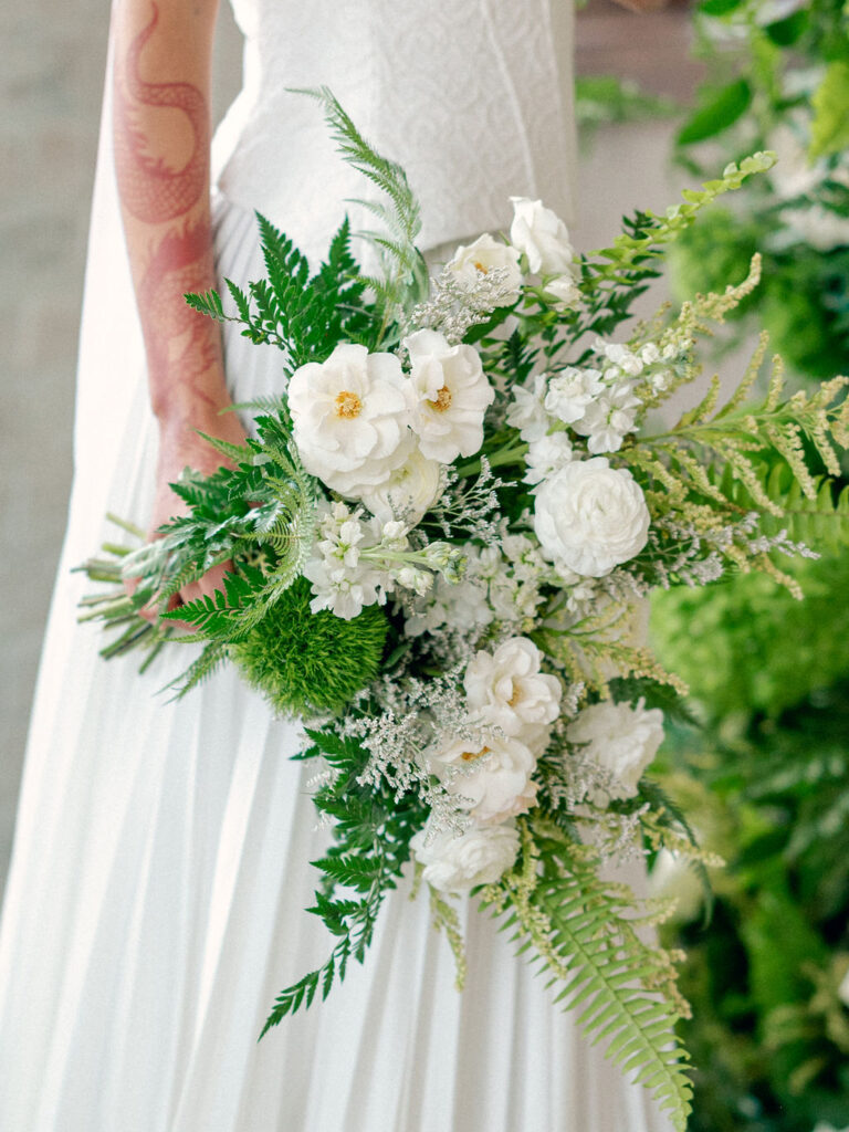 A bride in a drop waist wedding gown holds a wedding bouquet of greenery and white florals.