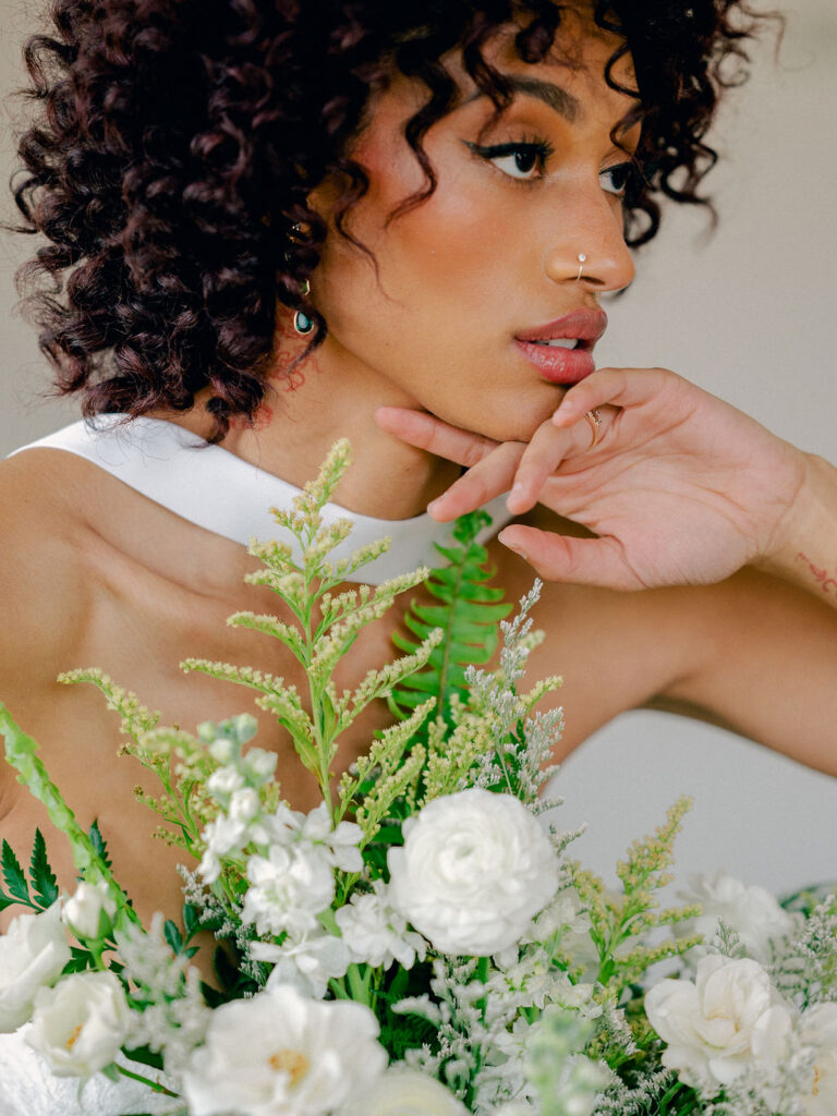 A bride in a drop waist wedding gown with a neck scarf draped behind her holds a bouquet inspired by ferns with lush greenery and white florals.