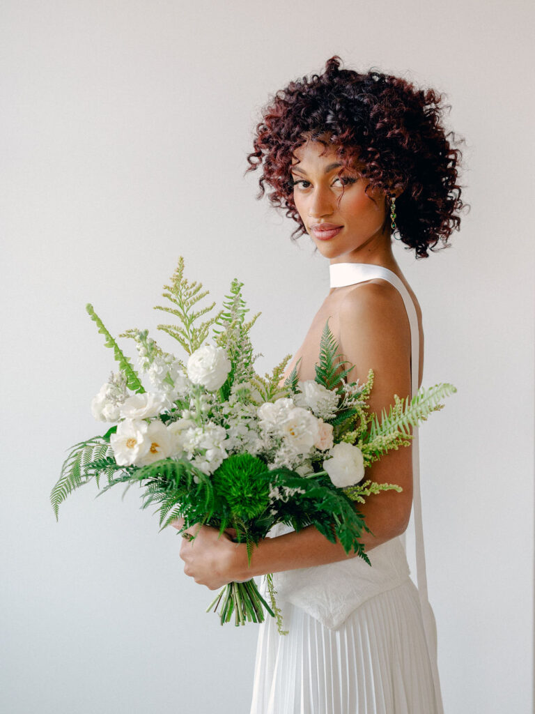 A bride stands in front of a white wall in a high end drop waist wedding gown with a neck scarf draped down her back. She is holding a fern-inspired bouquet of greenery and white flowers.