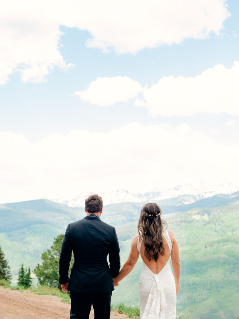 A bride and groom walk together on Vail Mountain, looking out at a stunning blue sky summer day.