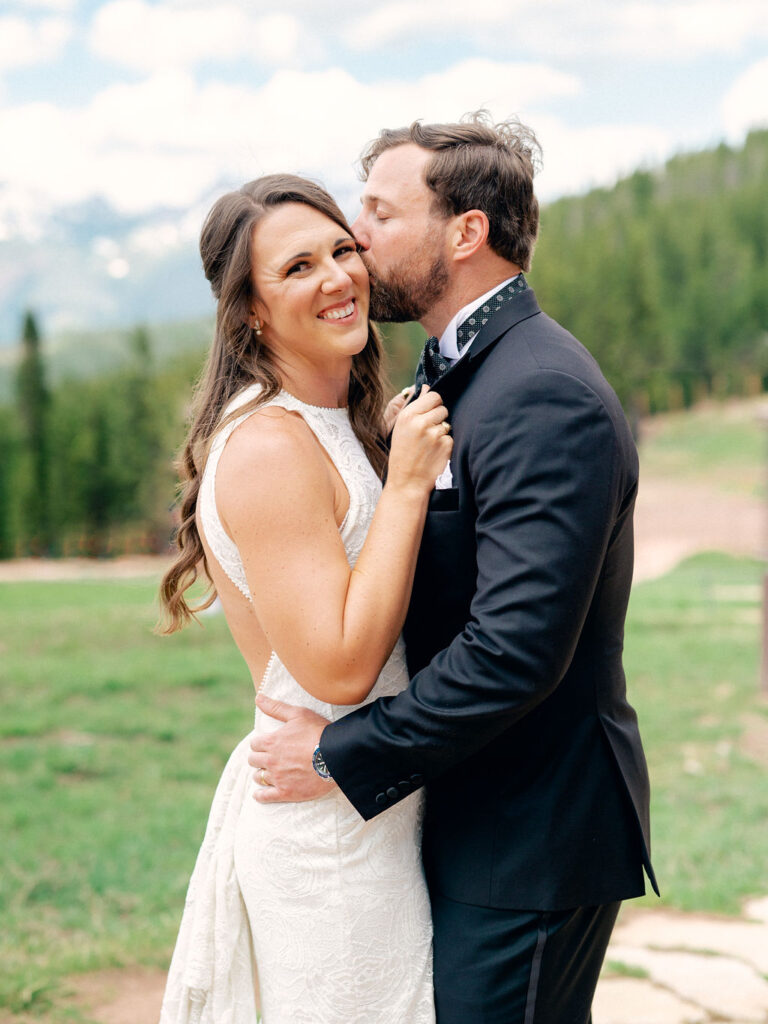 A groom kisses his bride as she smiles at the camera in a sweet wedding portrait in Vail, Colorado.