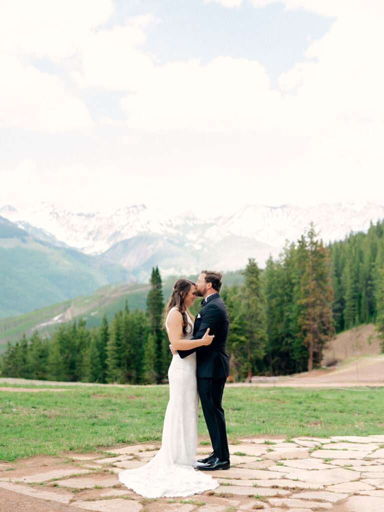 Standing among the mountains of Vail Colorado, a groom kisses his bride's forehead on a colorful summer day.