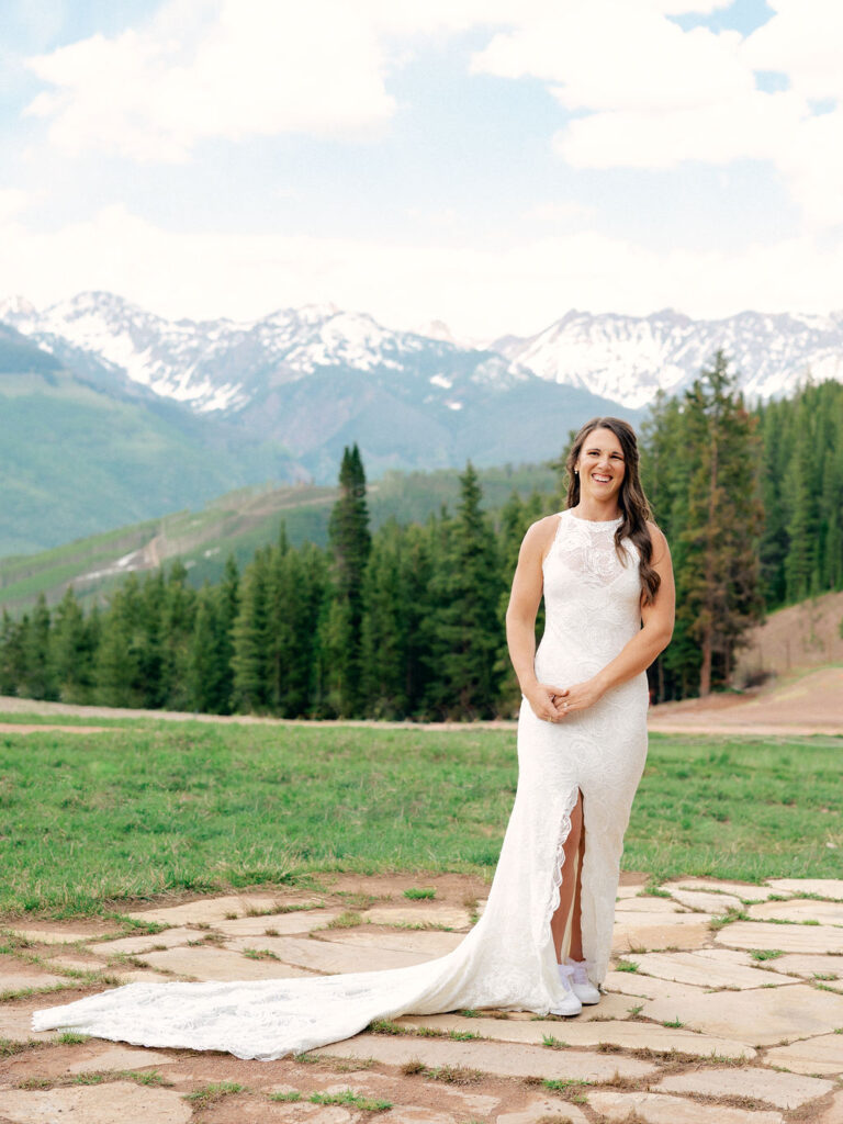 A bride in a high neck lace gown with a long train smiles for a classic portrait on top of Vail Mountain.