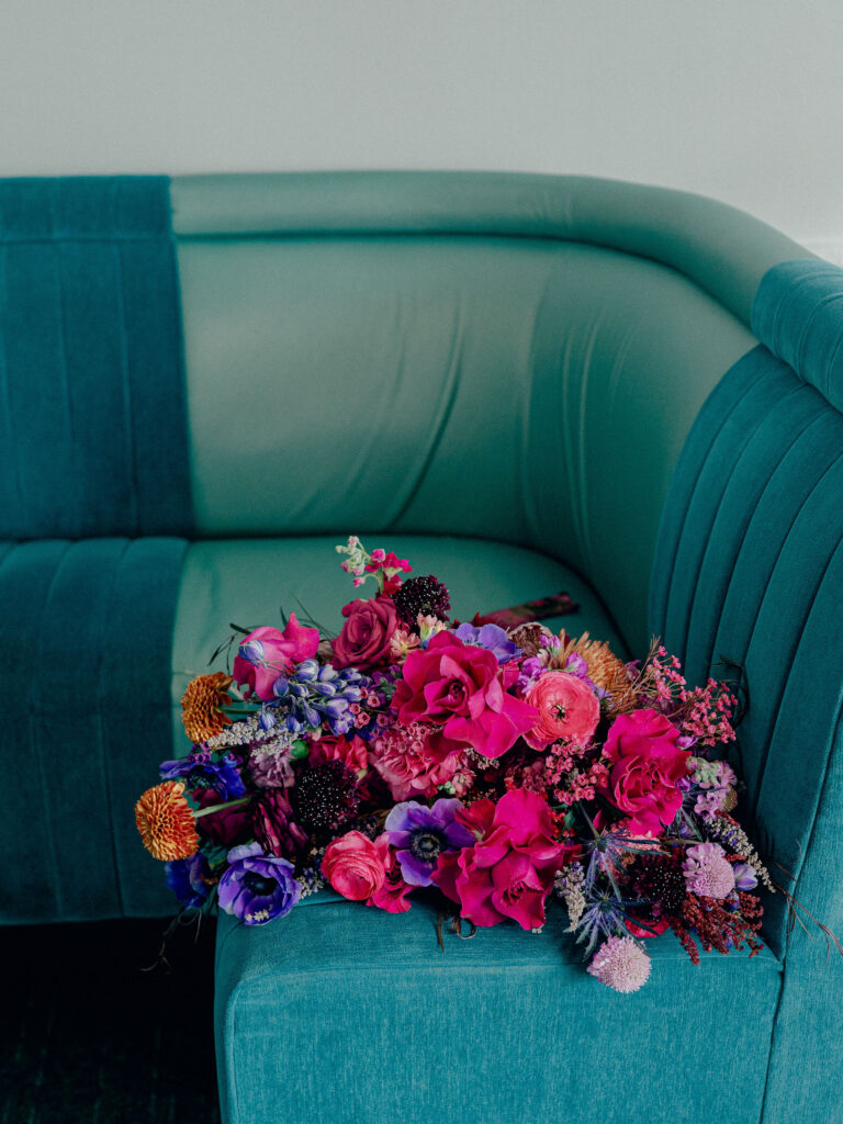 A bouquet of vibrant jewel tone florals sits on a teal couch. The wedding florals included magenta roses, purple anenomes, pink ranunculas, and more.