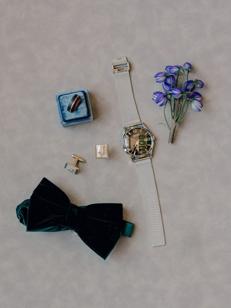 A detail flatlay of a groom's wedding day details, including a green velvet bow tie, watch, wedding ring, and custom made cufflinks.