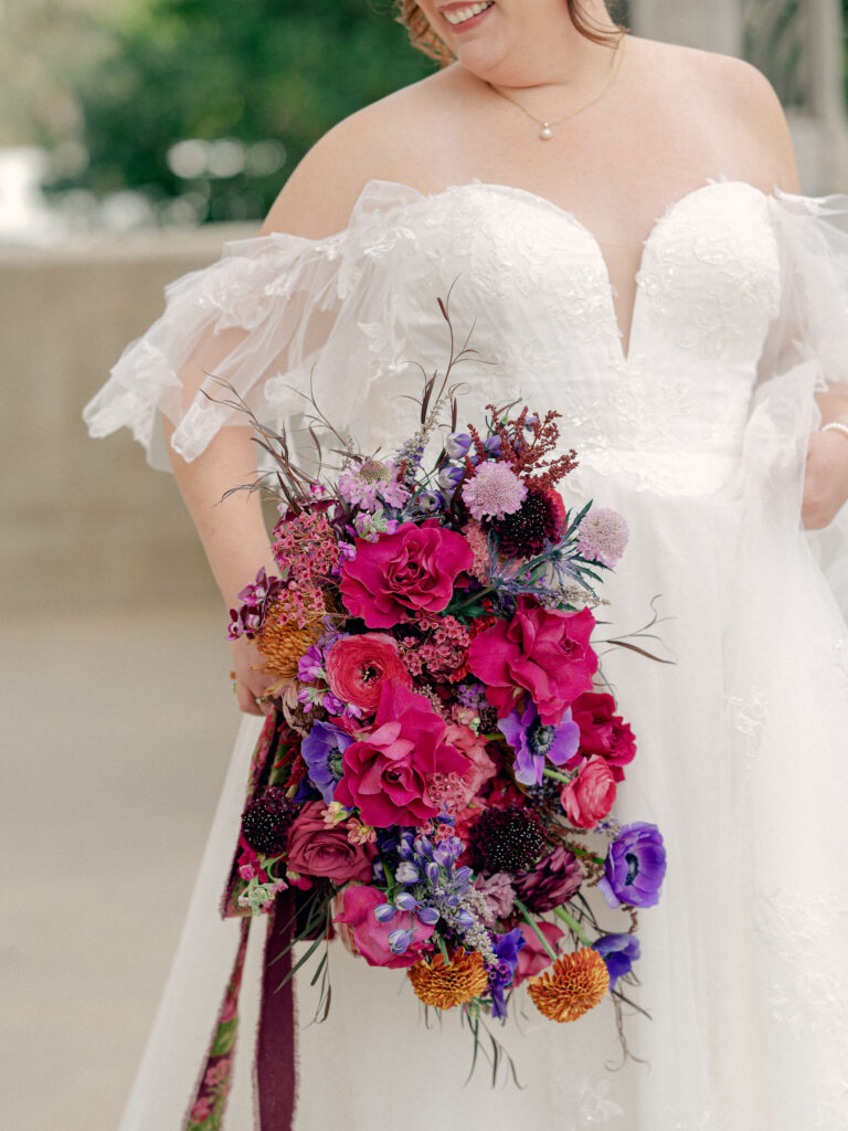 A bride in a lace wedding gown with off the shoulder sleeves holds her large bouquet of jewel tone flowers at her Oklahoma City wedding.