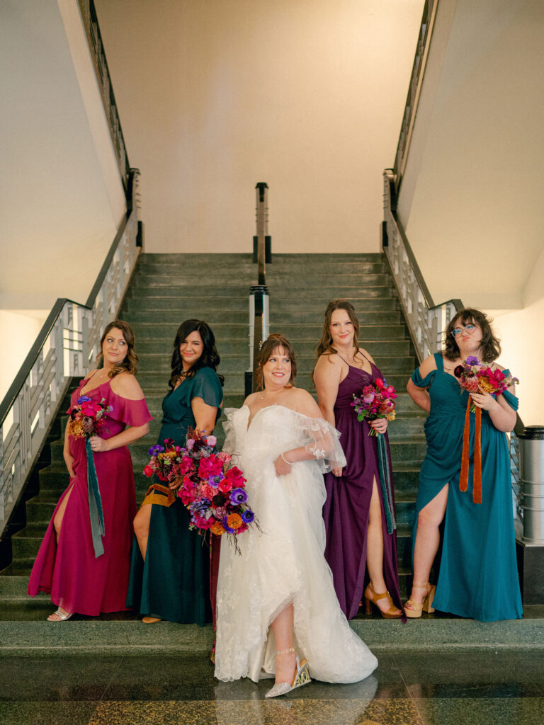 A bride and her bridesmaids dressed in jewel tone colors pose on the stairs of her historic Oklahoma City wedding venue, the OKC Civic Center.