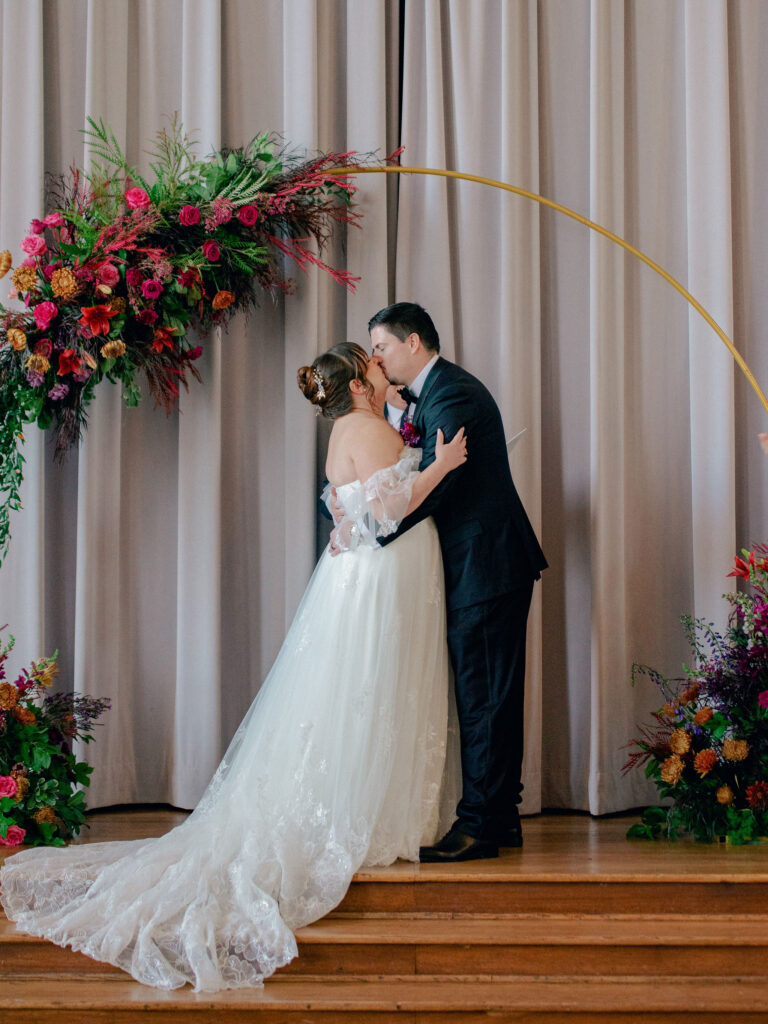 A bride and groom kiss under an arch of jewel tone florals at their Oklahoma City wedding.