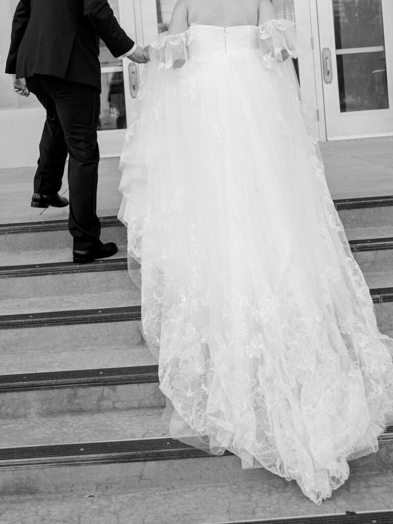 A bride and groom walk up the stairs of the OKC Civic Center with her lace train trailing behind her.