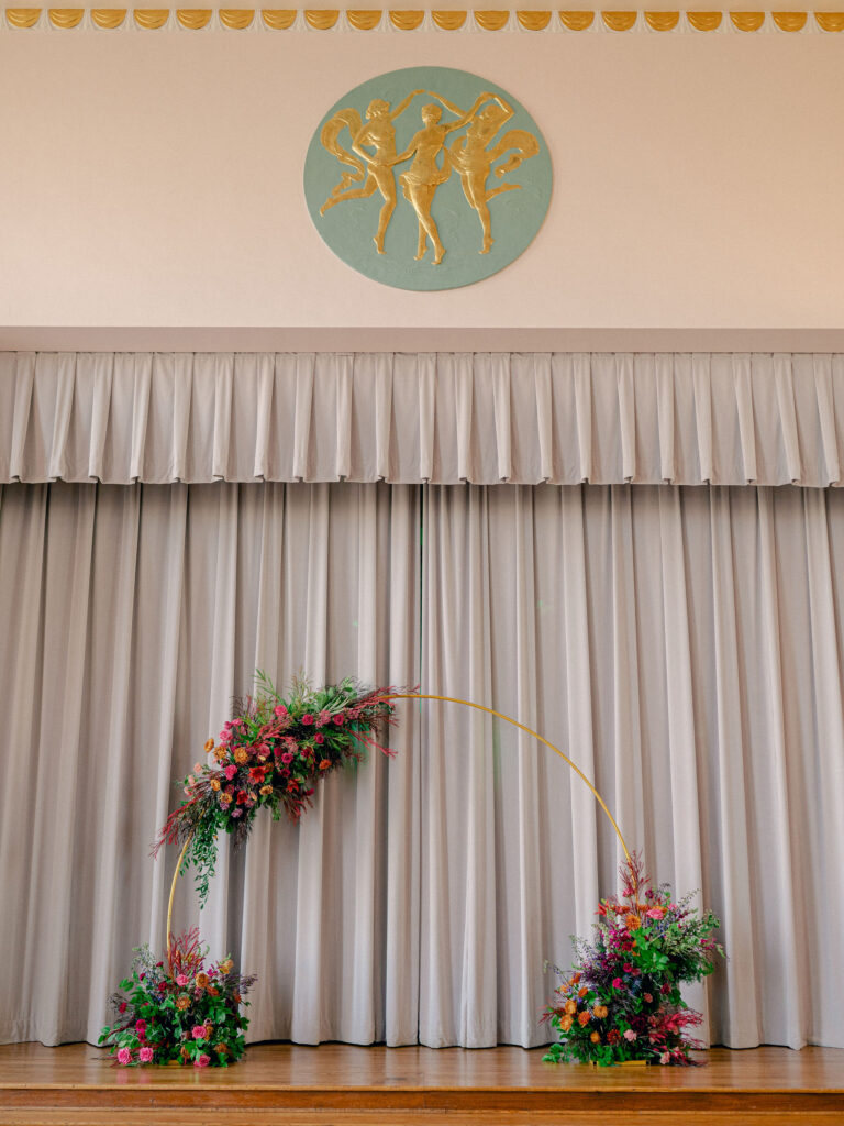 A gold arch covered in jewel tone florals sits on the stage of an art deco wedding venue for an Oklahoma City wedding ceremony.