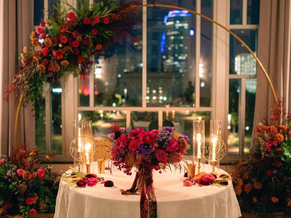 The sweetheart table at this Oklahoma City wedding reception was framed by an arch of jewel tone florals in front of a window that looked out on Downtown OKC.