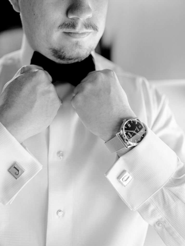 A groom straightens his velvet bow tie. He is wearing a watch and custom make pearl and gold cufflinks.